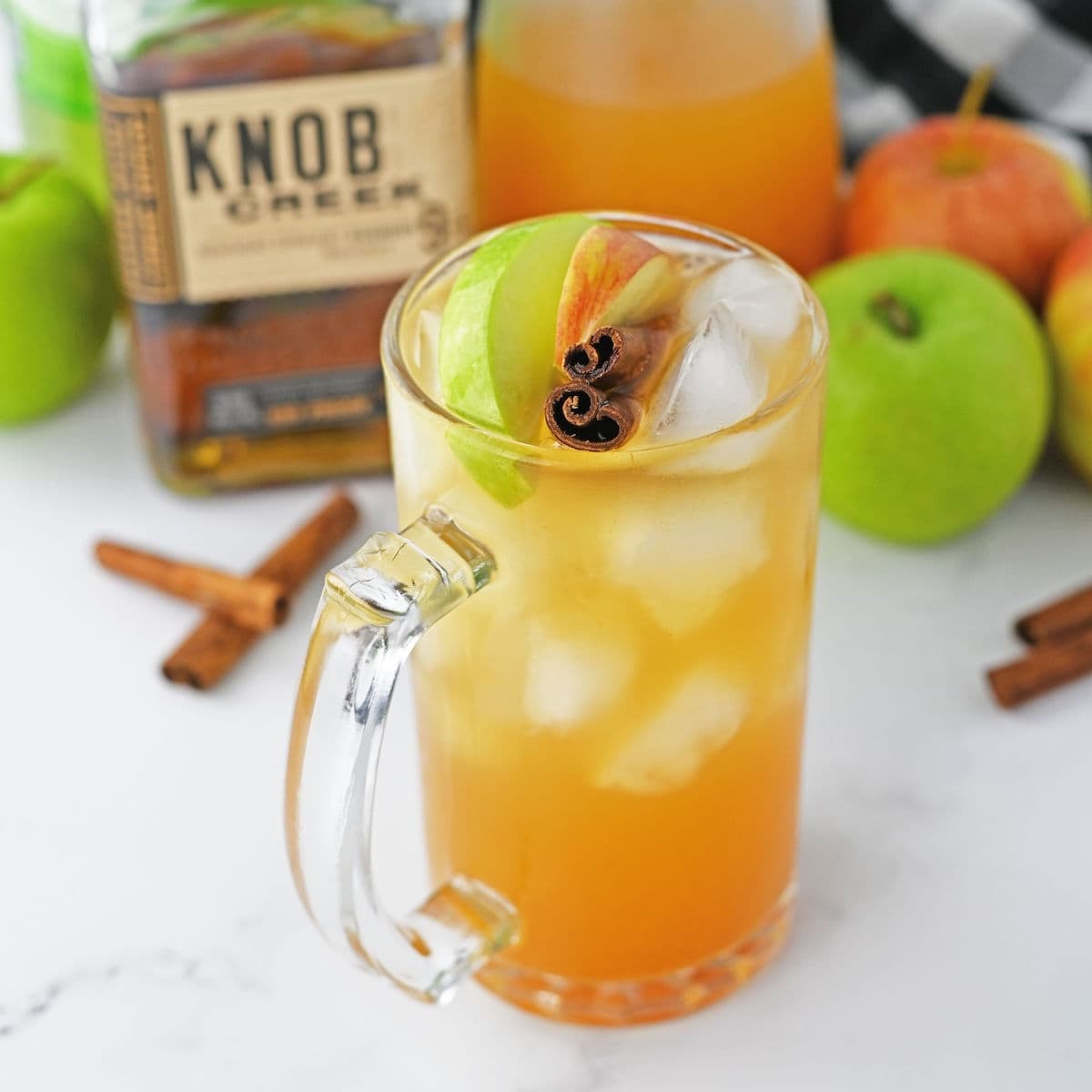 Glass of apple cider with cinnamon stick and apple slices with bourbon bottle and apples and ginger ale in background.