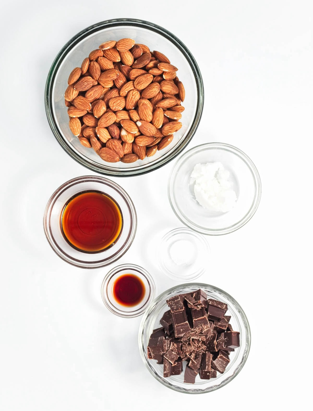 Glass bowls of almonds, maple syrup, oil, salt, vanilla, and chocolate.