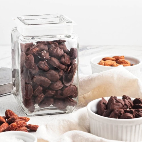 Jar of chocolate covered almonds with more in a bowl on a white napkin with a jar of more behind and a small bowl of raw almonds in background.