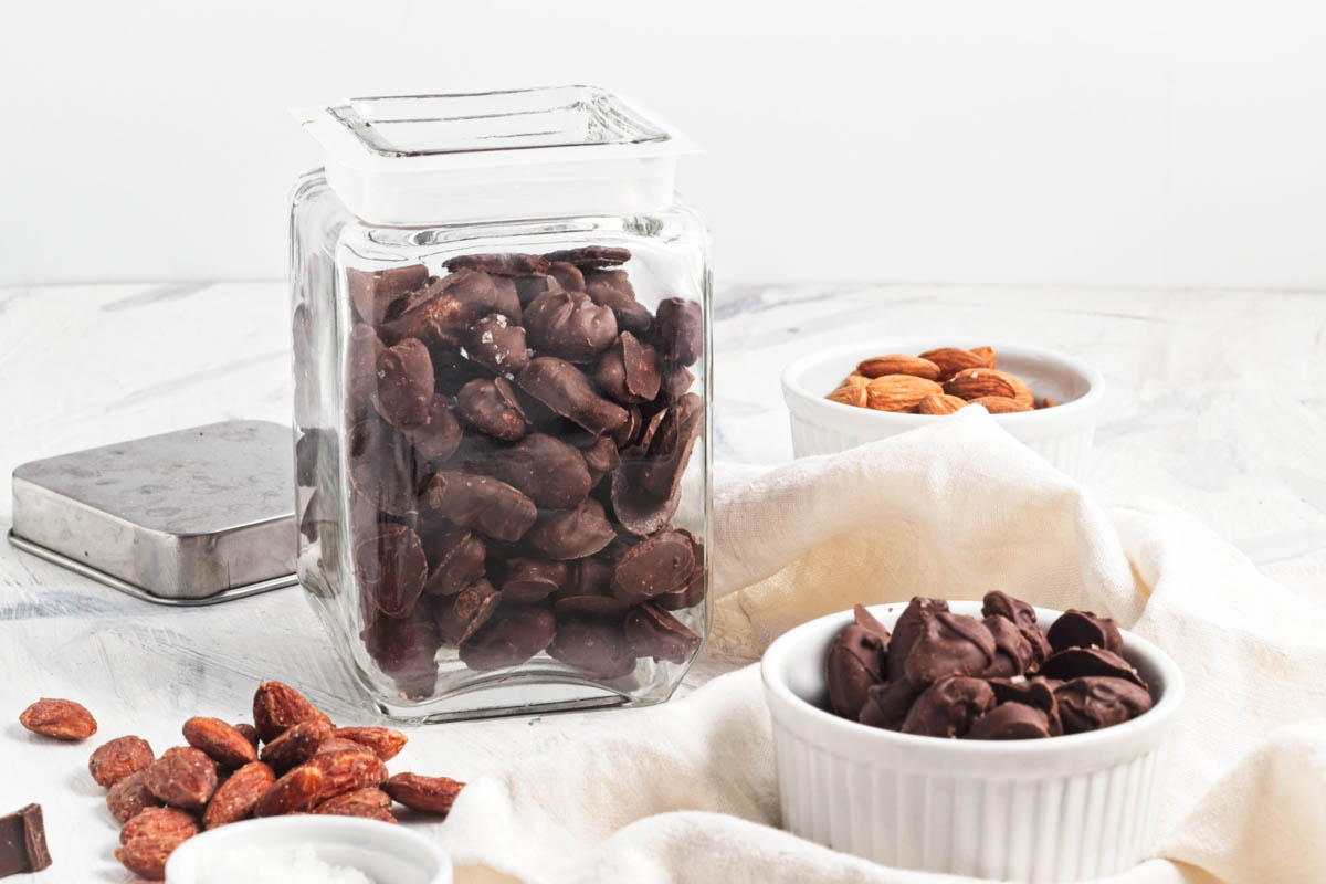 Jar of chocolate covered almonds with more in a bowl on a white napkin with a jar of more behind and a small bowl of raw almonds in background.