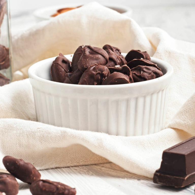 Chocolate covered almonds in a bowl on a shite napkin with a jar of more behind and a small bowl of raw almonds in background.