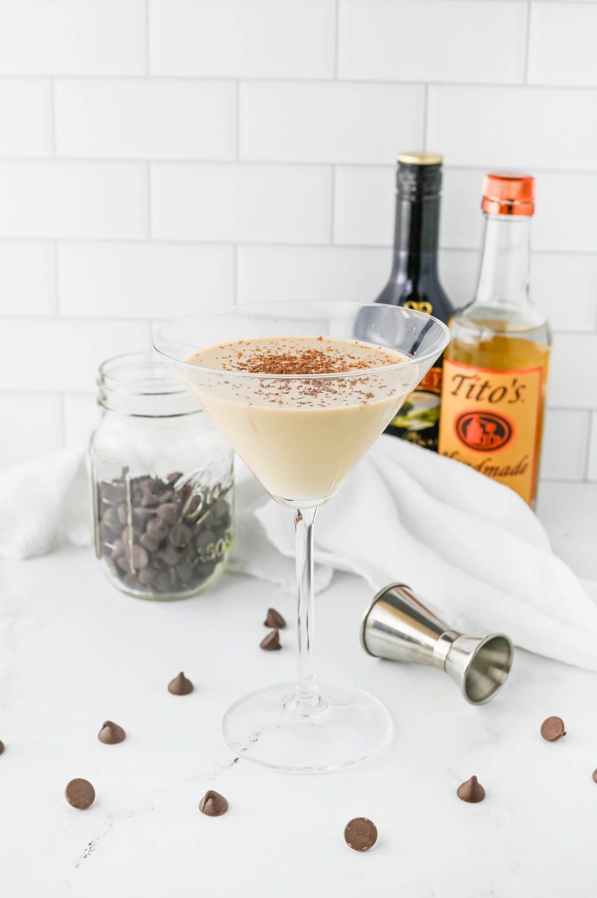 Creamy cocktail in a martini glass in front of white napkin with chocolate chips in jar and scattered and an aluminum shot container on table and bottles in background.