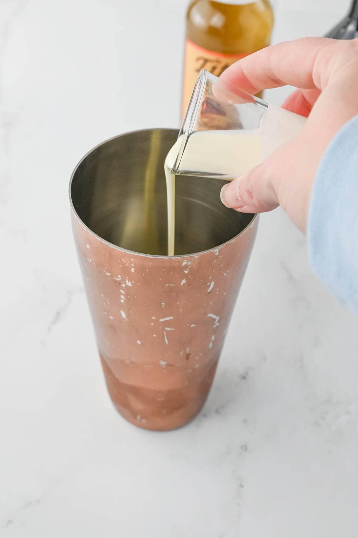 Pouring cream from a shot glass into a shaker with a bottle of liquor in background.