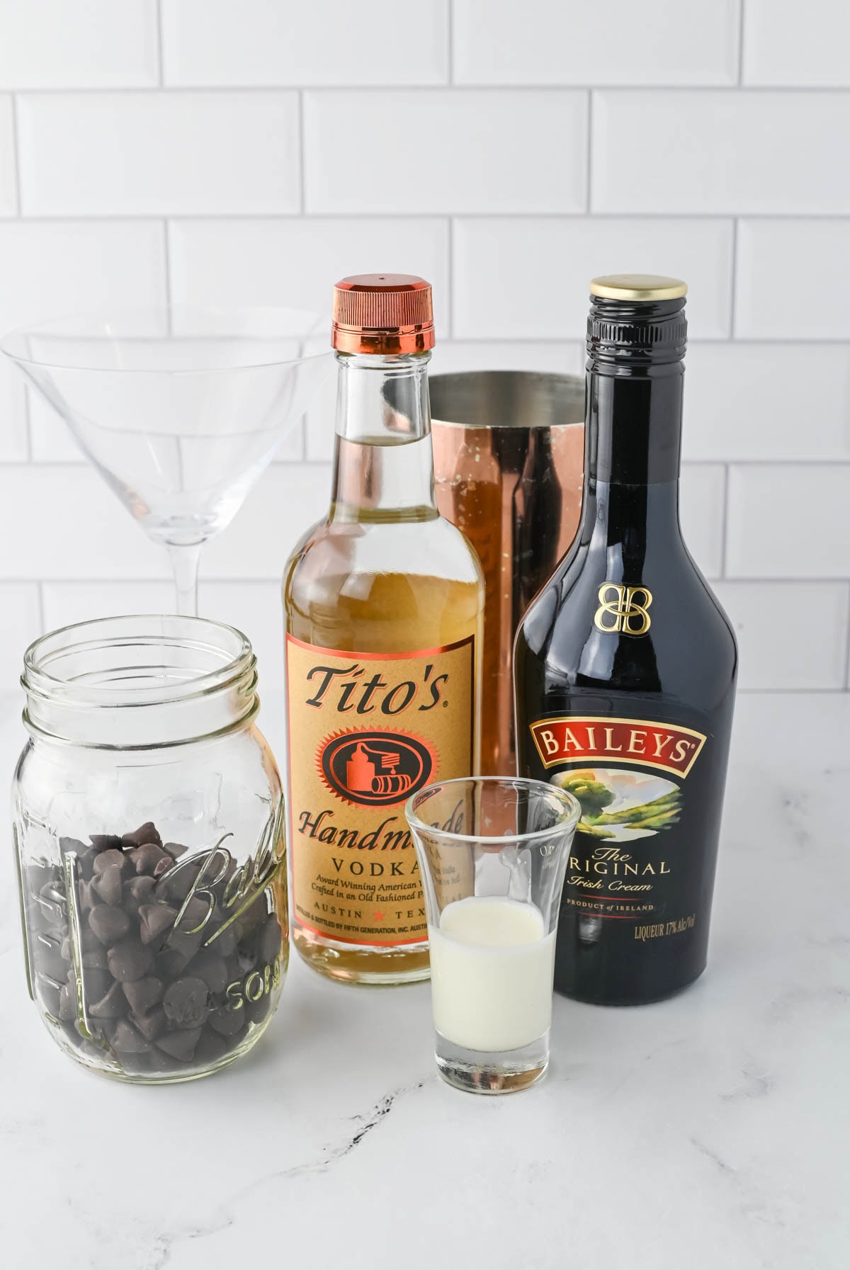 Bottles of Baileys and Titos vodka in front of a cocktail shaker with jar of chocolate chips and shot glass of cream in front.