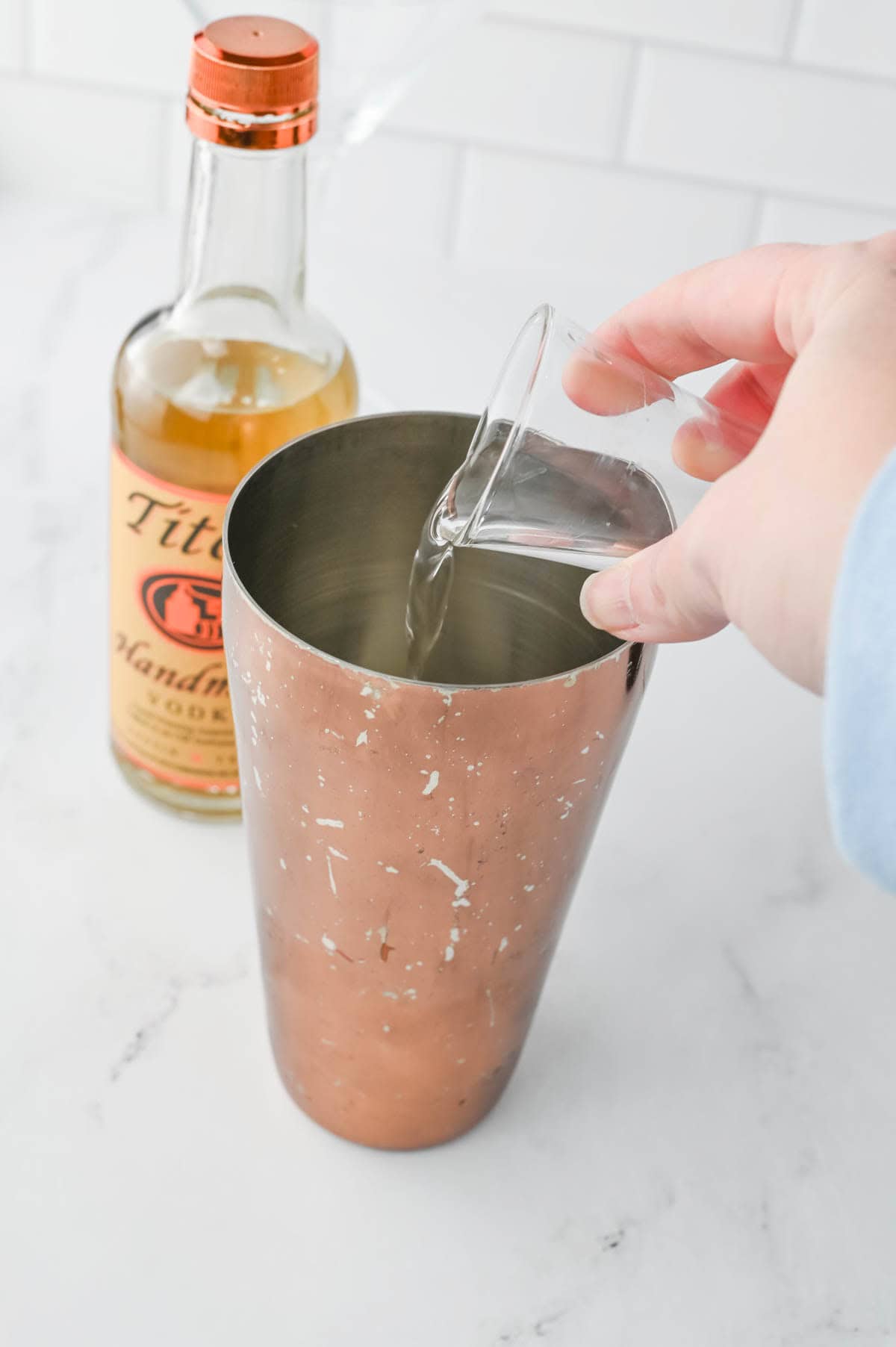 Pouring clear liquid into a shaker glass with bottle in background.