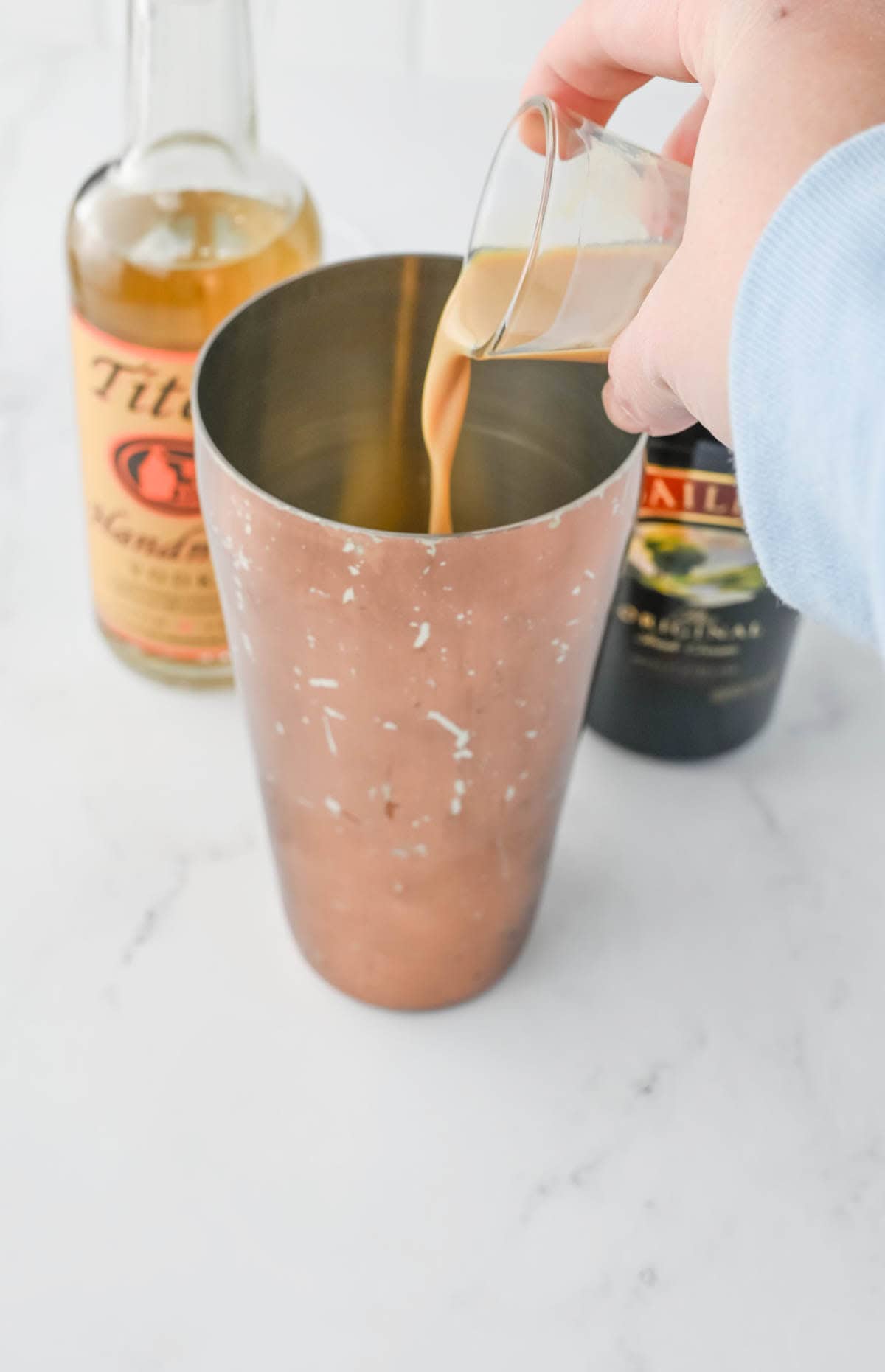 Pouring Irish cream into a cocktail shaker with bottles in background.