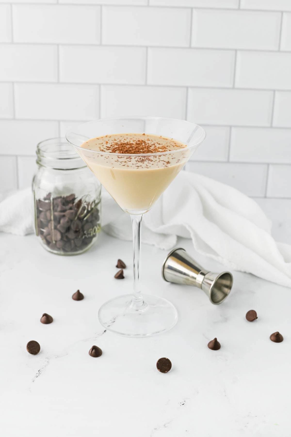 Creamy cocktail in a martini glass with chocolate chips in jar and scattered and an aluminum shot container on table.