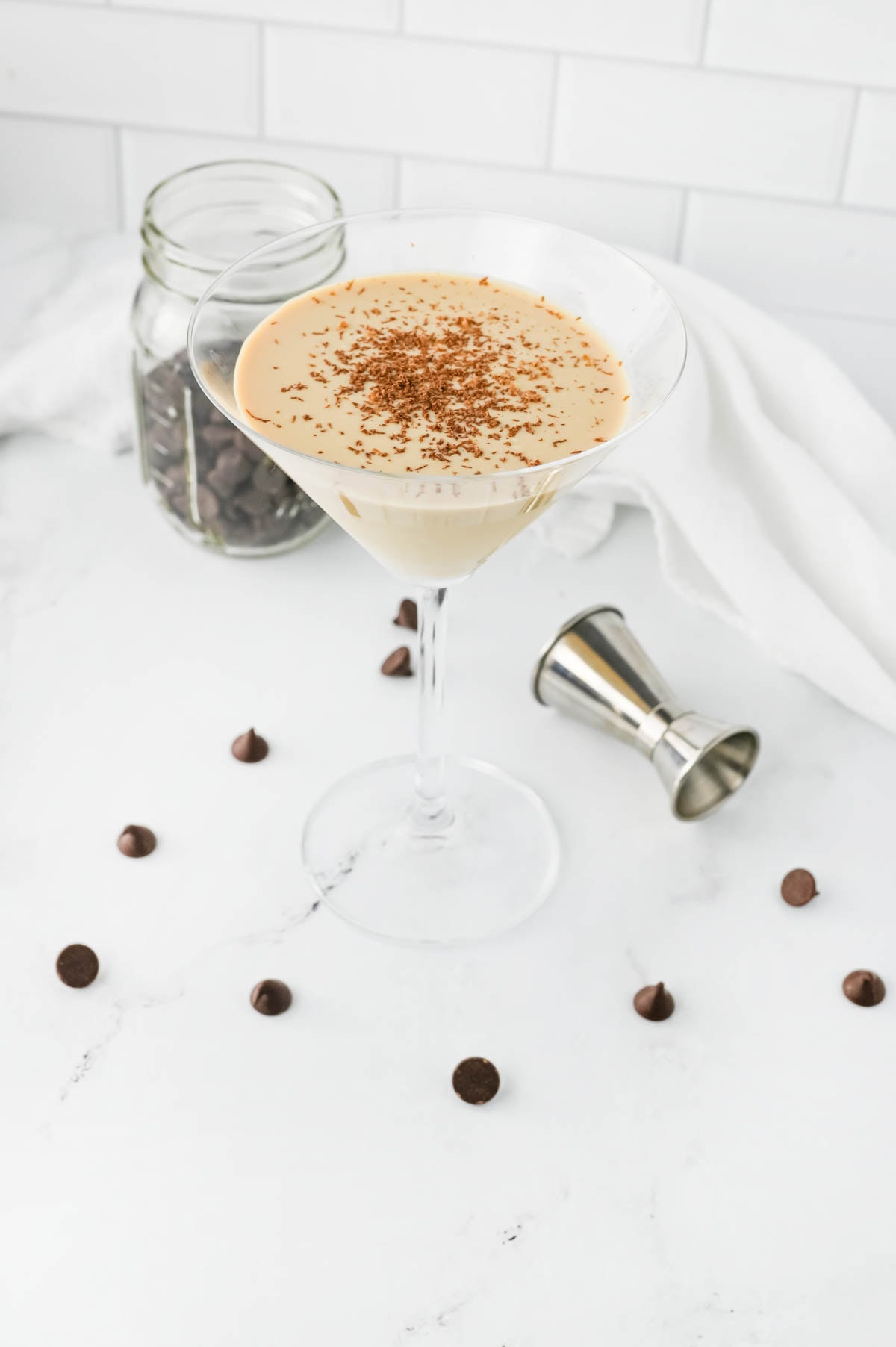 Creamy cocktail in a martini glass with chocolate chips in jar and scattered and an aluminum shot container on table.