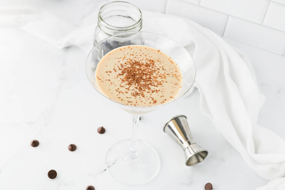 Creamy cocktail in a martini glass with chocolate chips in jar and scattered and an aluminum shot container on table with white napkin in background.