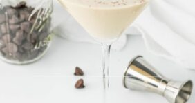 Creamy cocktail in a martini glass with chocolate chips in jar and scattered and an aluminum shot container on table image for Pinterest.