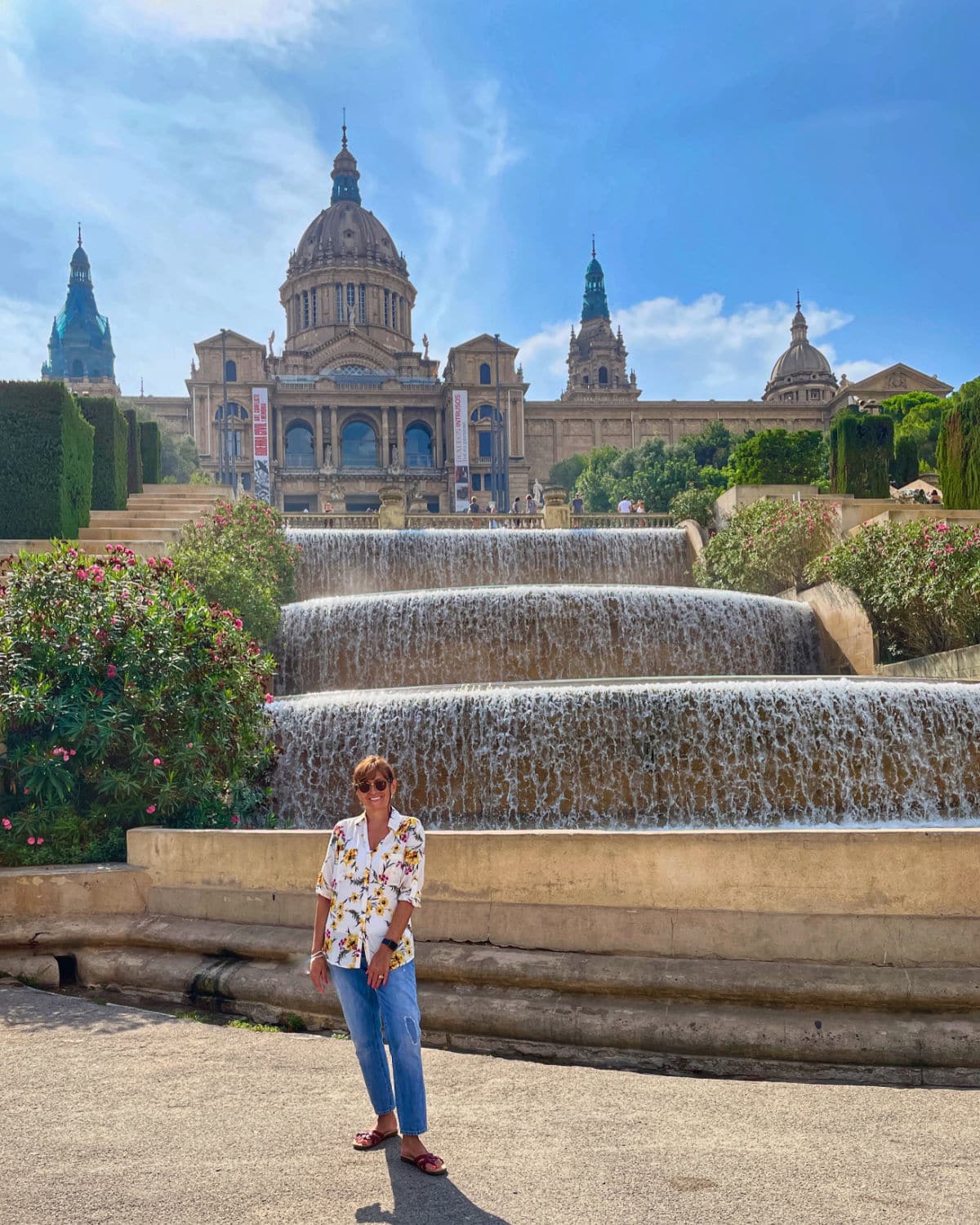 Woman standing in front of large fountain with a large historic building in background against a blue sky.