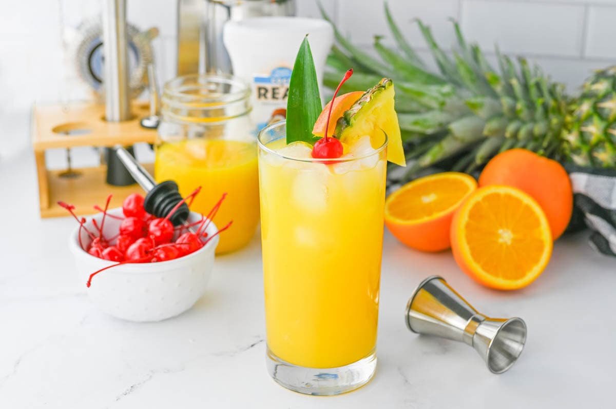 Orange colored cocktail with cherry and pineapple slice in a tall glass on a white counter with ingredients in background.