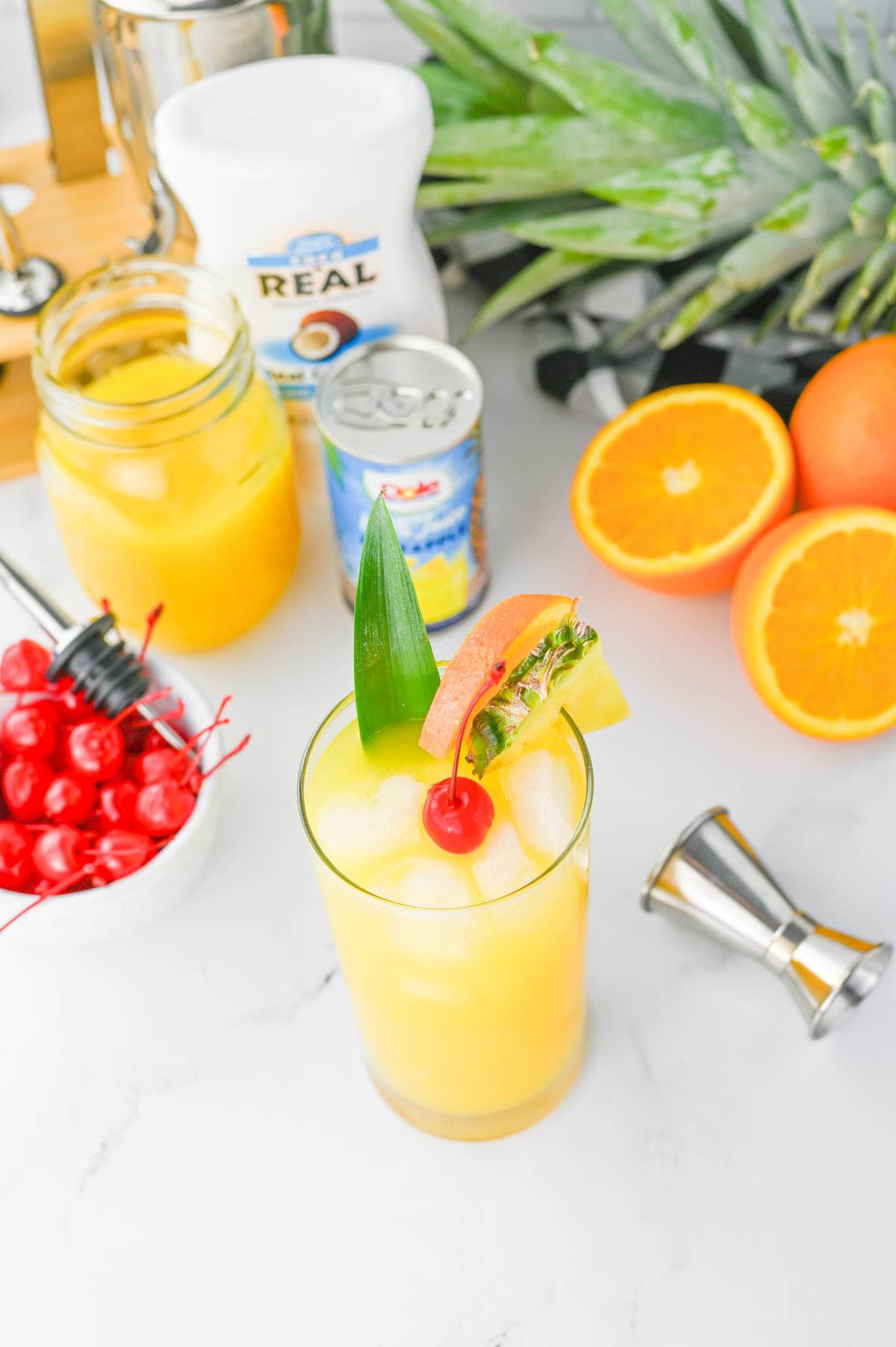 Orange colored cocktail with cherry and pineapple slice in a tall glass and silver shot glass on a white counter with ingredients in background.