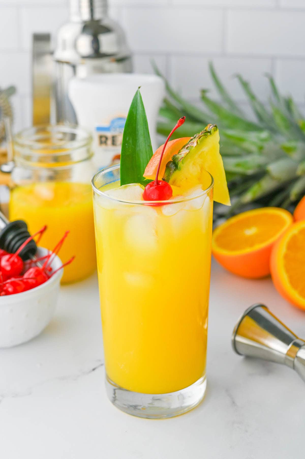 Orange colored cocktail with cherry and pineapple slice in a tall glass on a white counter with ingredients in background.