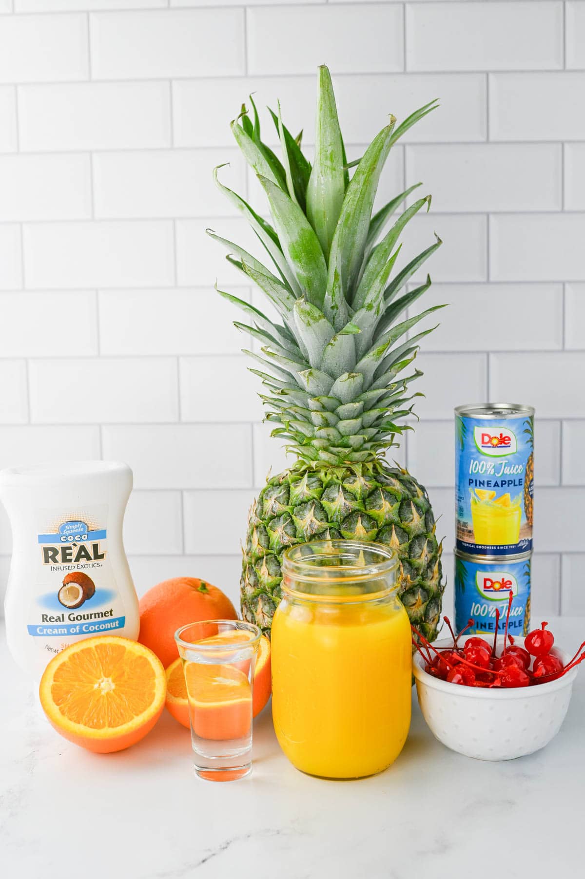 Mason jar of orange juice, shot of rum, fresh orange, coconut cream, fresh pineapple, pineapple juice, and maraschino cherries on a white counter.