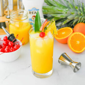 Orange colored cocktail with cherry and pineapple slice in a tall glass on a white counter with ingredients in background.