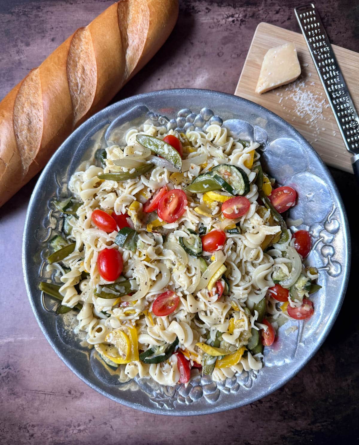 Pasta and vegetables with seasoning and parmesan cheese with a block of cheese and grater and a baguette to the side.