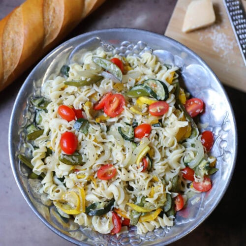 Pasta and vegetables with seasoning and parmesan cheese with a block of cheese and grater and a baguette to the side.