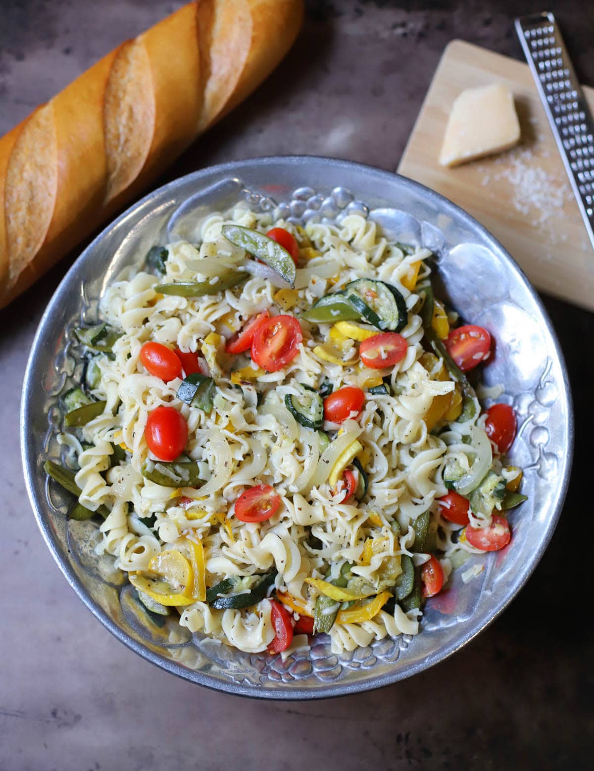 Pasta and vegetables with seasoning and parmesan cheese with a block of cheese and grater and a baguette to the side.