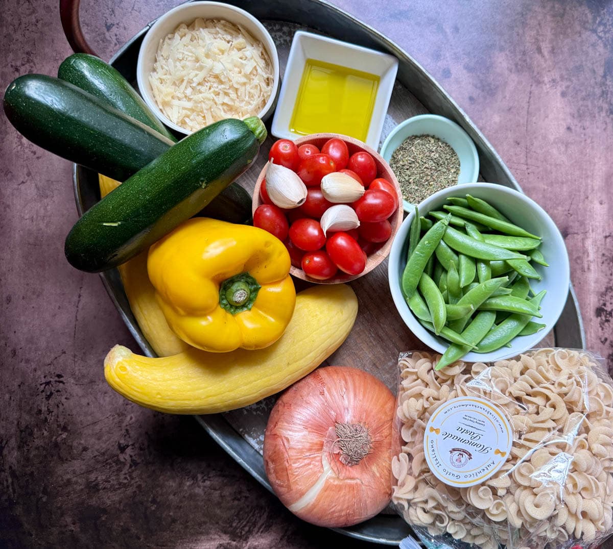 Fresh vegetables on a tray with a bog of pasta, bowl of parmesan cheese, bowl of olive oil, and bowl of spices.