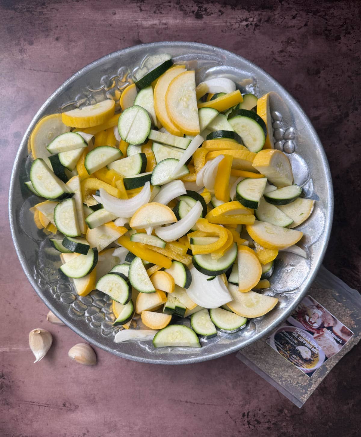 Fresh yellow squash and zucchini with onions in silver bowl and cloves of garlic with a seasoning pack on table.