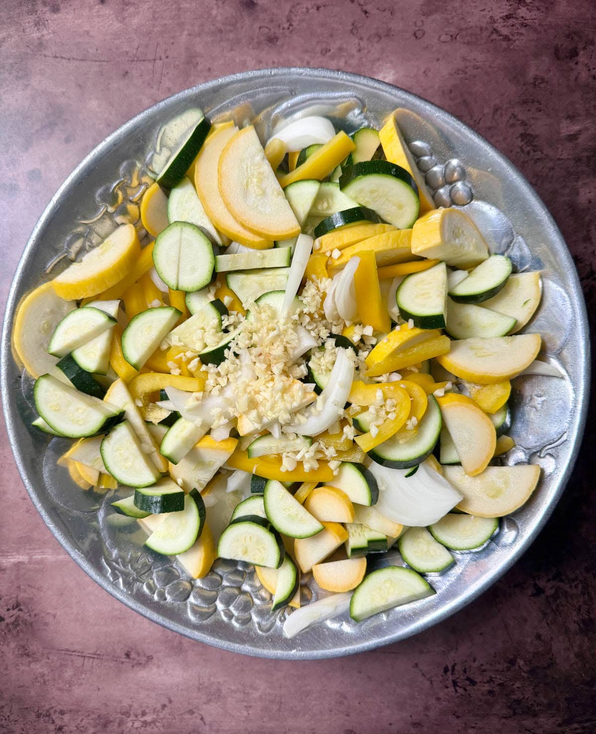 Fresh yellow squash and zucchini with onions and chopped garlic in silver bowl on table.