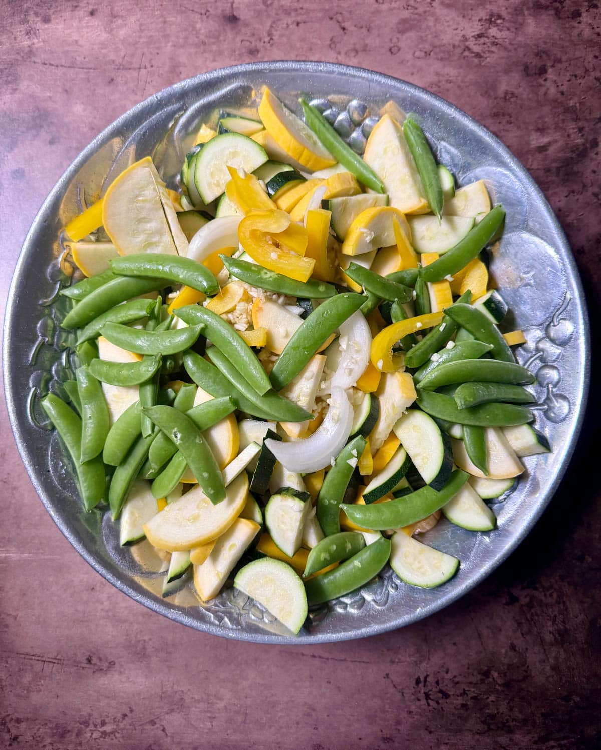 Fresh yellow squash, zucchini, and snap peas with onions and chopped garlic in silver bowl on table.