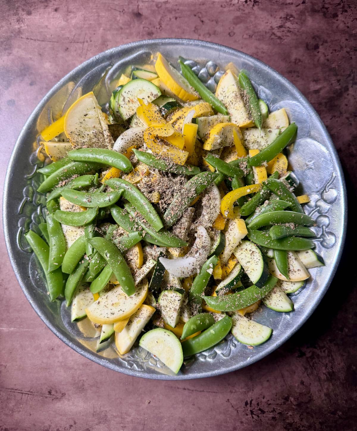 Fresh yellow squash, zucchini, and snap peas with onions and chopped garlic topped with dried seasoning in silver bowl on a table