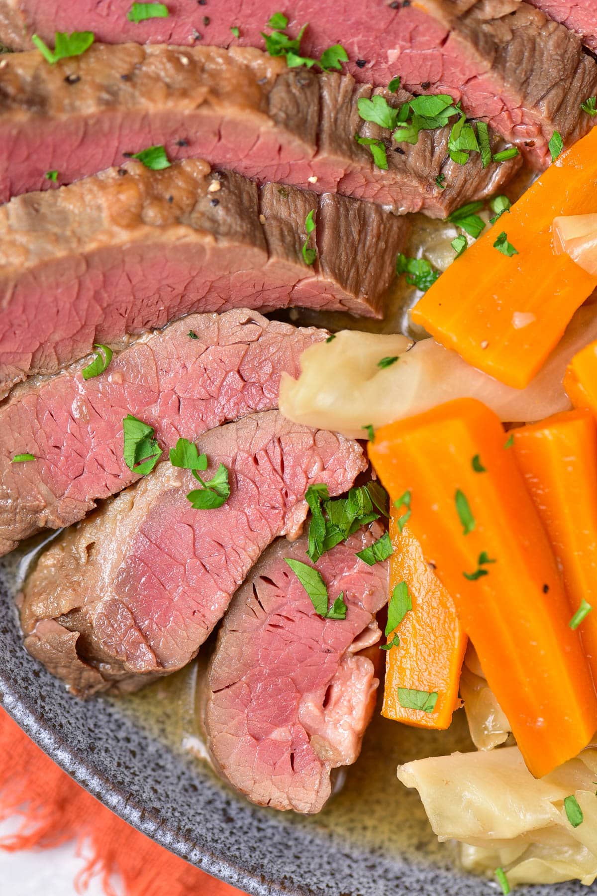 Close up sliced corned beef, carrots, cabbage, and potatoes on a grey plate over an orange napkin.