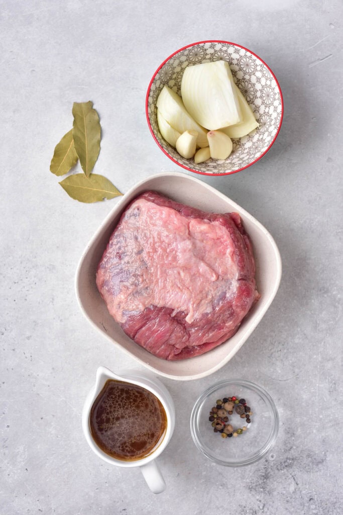 Bowl of corned bbef, bowl of onions and garlic, pitcher of broth, bowl of peppercorns, bay leaves to the side, on grey counter.