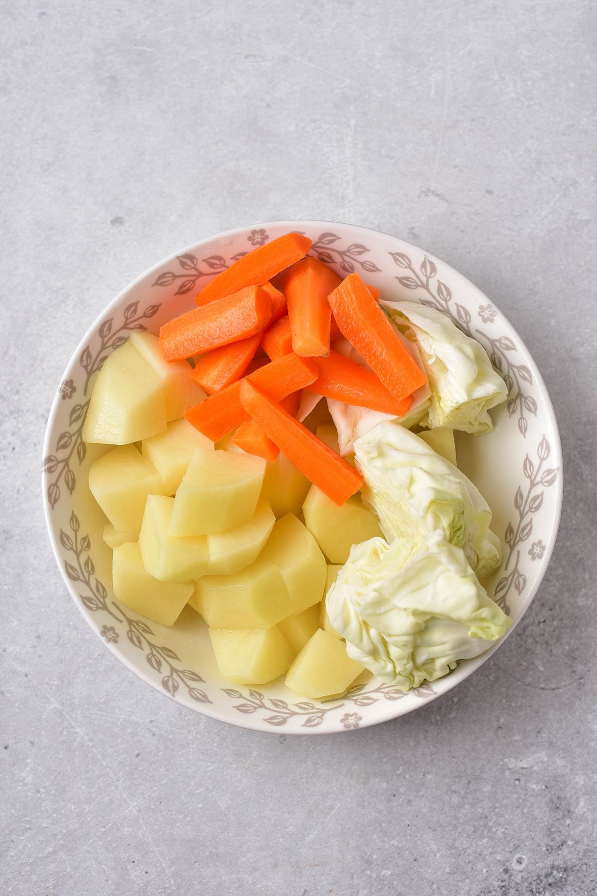 Sliced carrots, cubed potatoes, and cubed cabbage in a white bowl on a grey background.