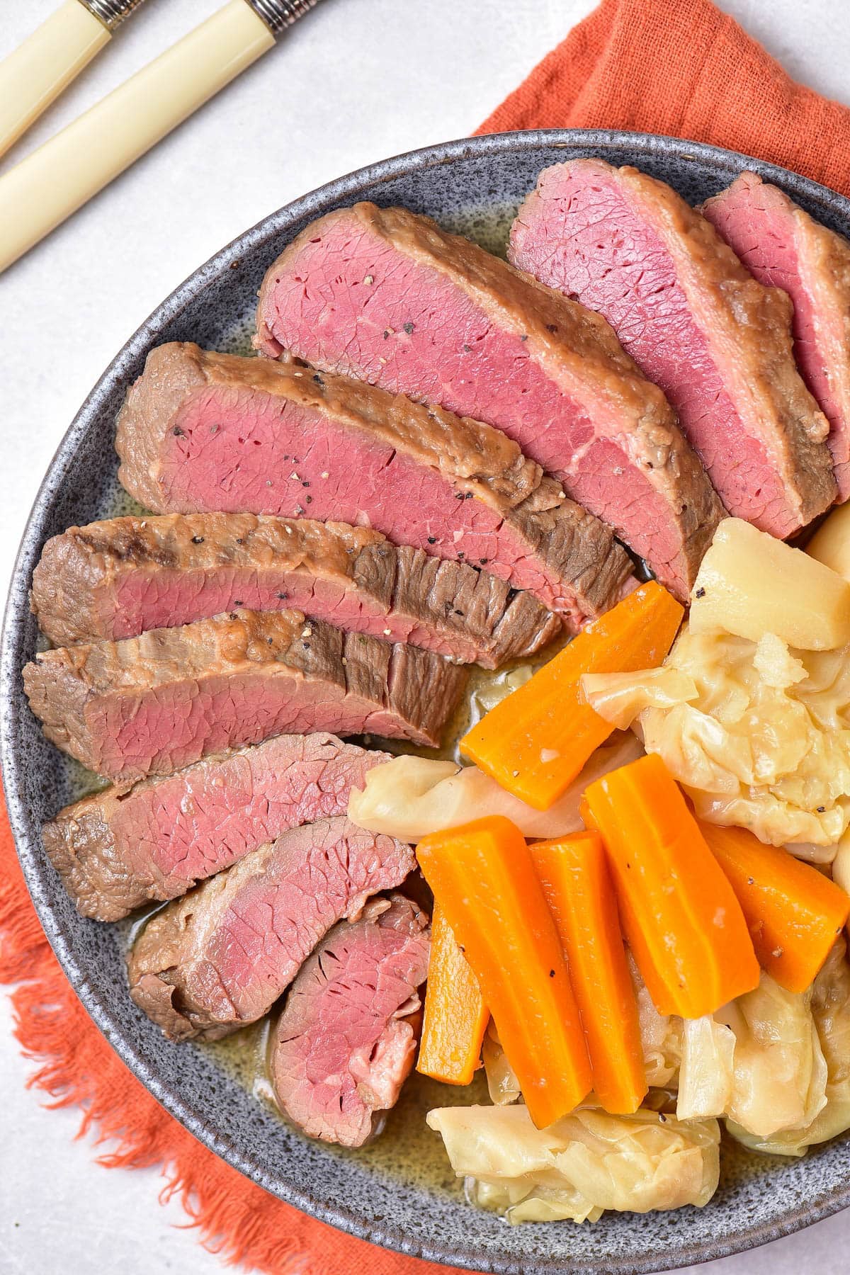 Sliced corned beef, carrots, cabbage, and potatoes on a grey plate over an orange napkin.