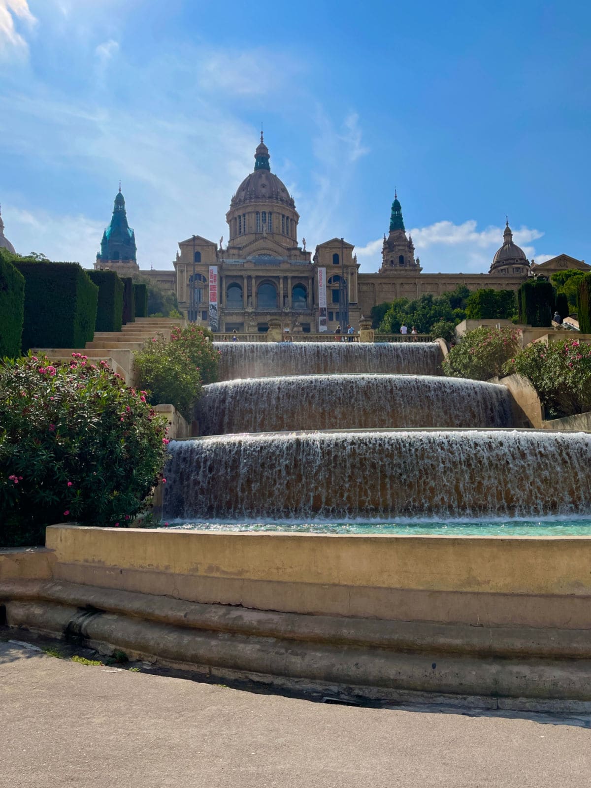 Impressive fountain in front of large historic building in background against a blue sky.