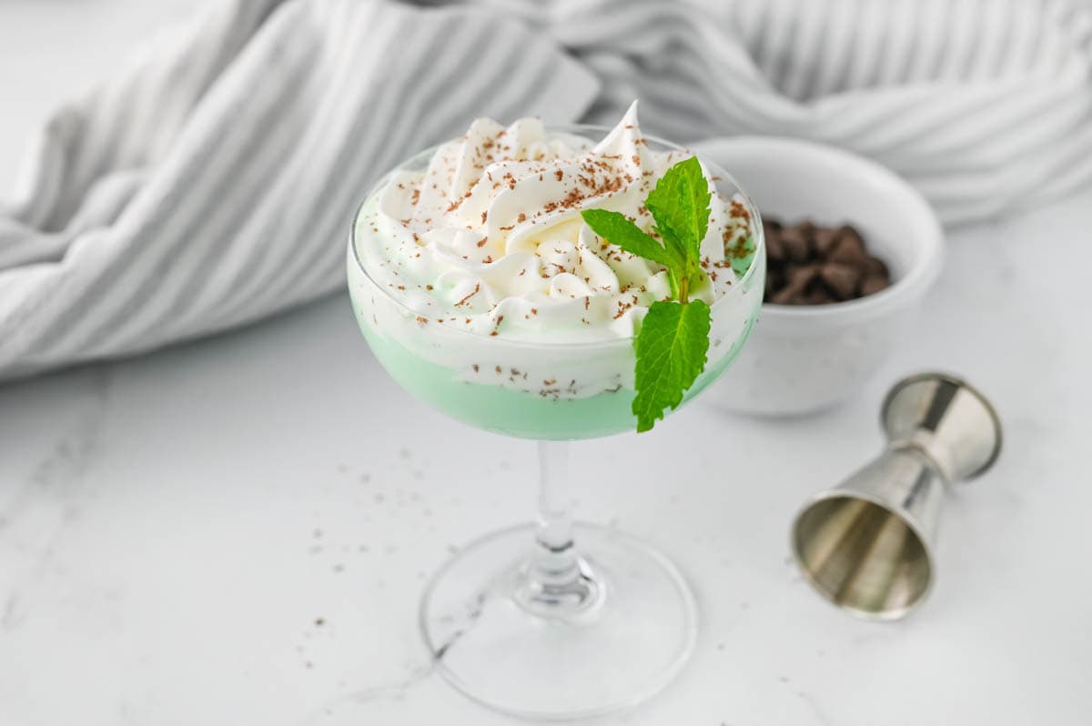 Green cocktail with whipped cream, chocolate shavings, a small bowl of chocolate chips, and a sprig of mint on marble counter with striped dish towel.