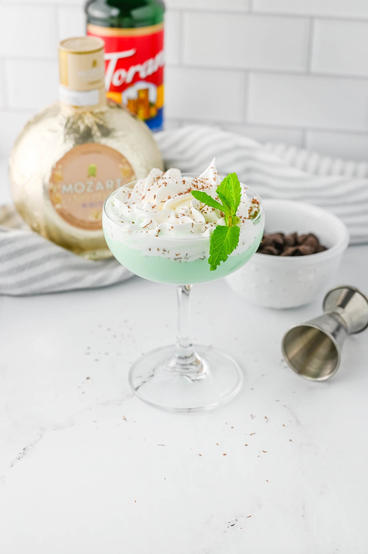 Green cocktail with whipped cream, chocolate shavings, a bowl of chocolate chips, a sprig of mint, and two bottles of liquor in the background on marble counter with striped dish towel.