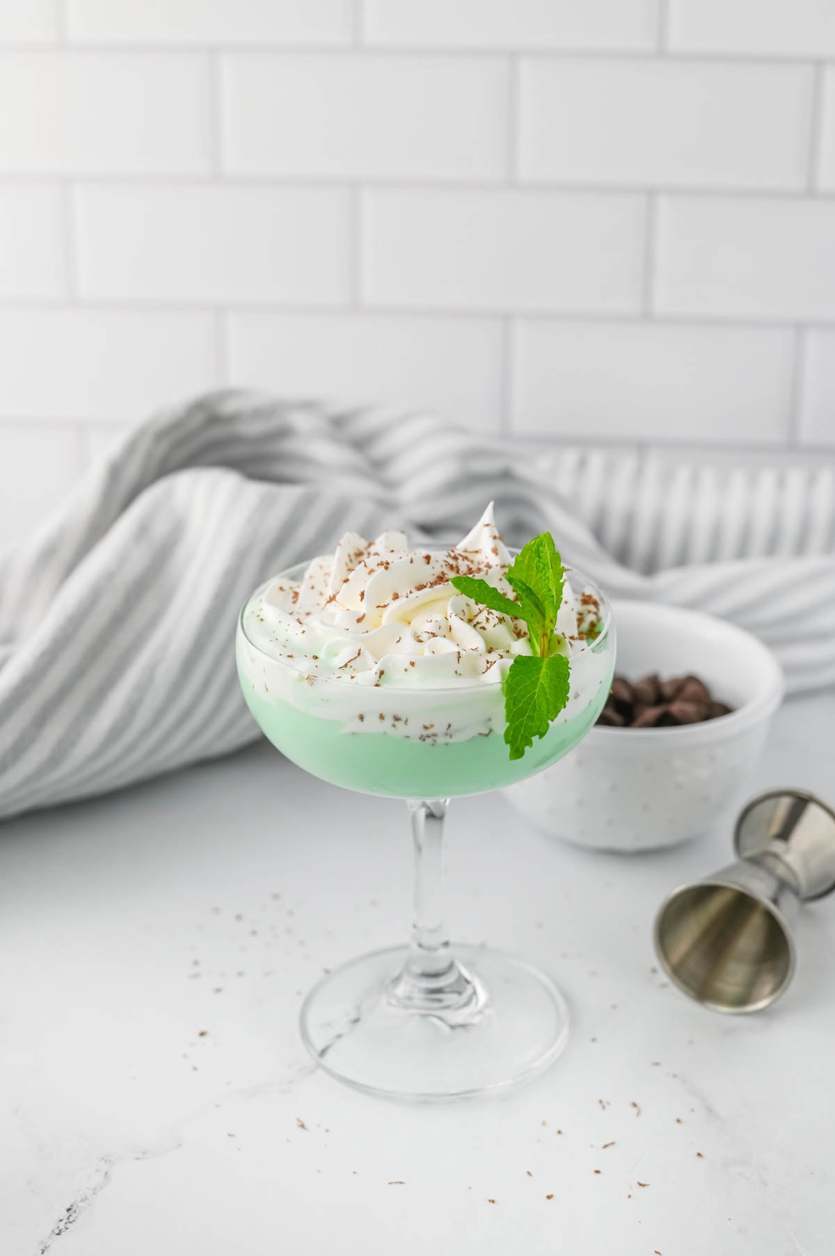 Green cocktail with whipped cream, chocolate shavings, and a sprig of mint on marble counter with striped dish towel.