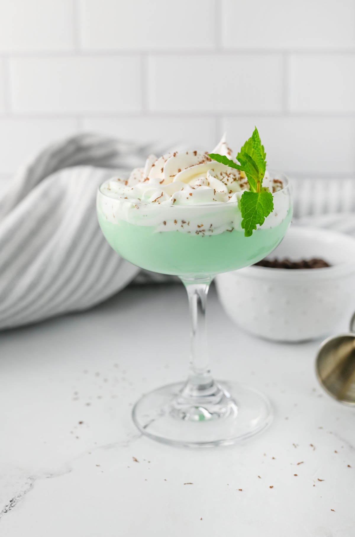 Green cocktail with whipped cream, chocolate shavings, and a sprig of mint on marble counter with striped dish towel.