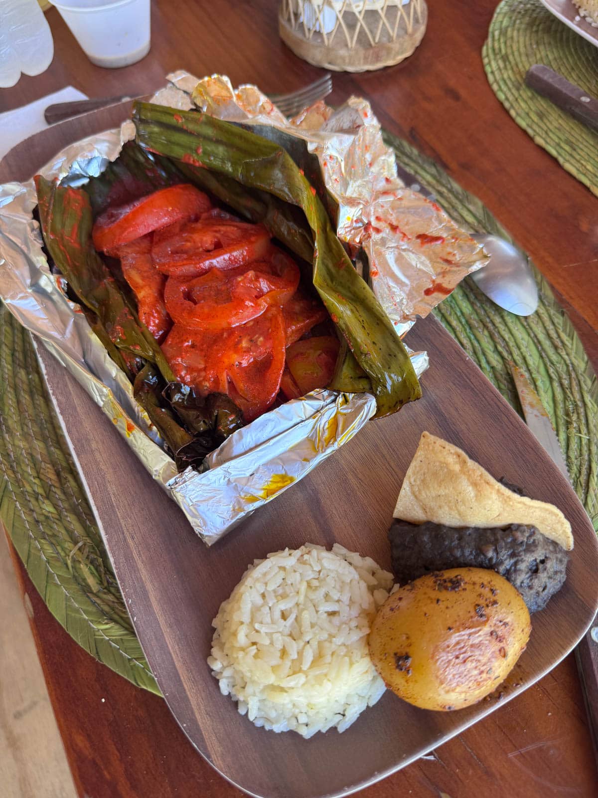 Plate with rice, potato, black beans and chip and aluminum wrapped vegetables in a banana leaf.