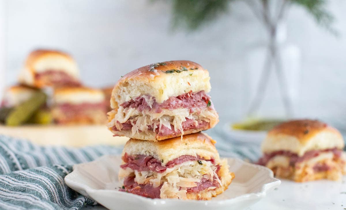 Small Reuben sandwiches on white plate with striped dish towel and more sandwiches in background.