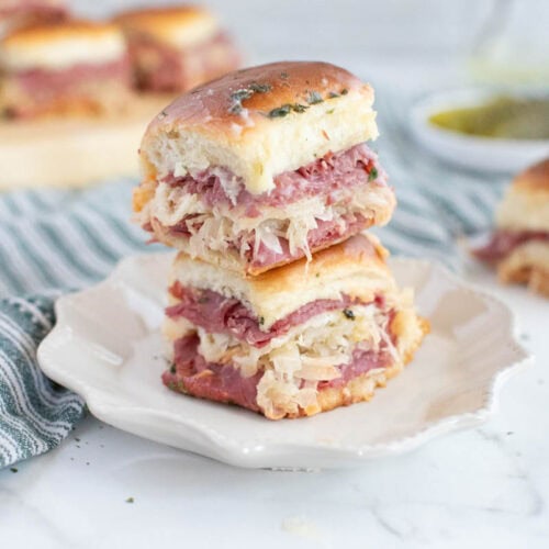 Small Reuben sandwiches on white plate with striped dish towel and more sandwiches in background.