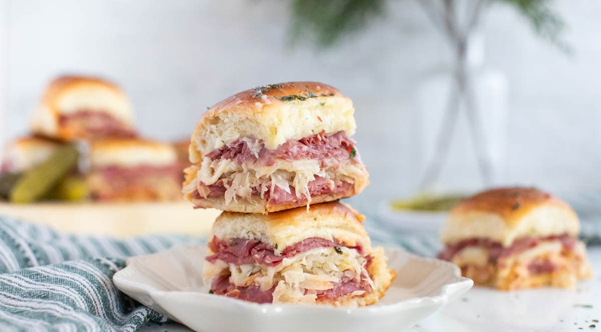 Small Reuben sandwiches on white plate with striped dish towel and more sandwiches in background.