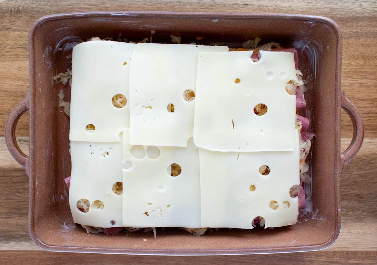 Sliced corned beef and sauerkraut topped with Swiss cheese on bottoms of rolls in a baking dish.