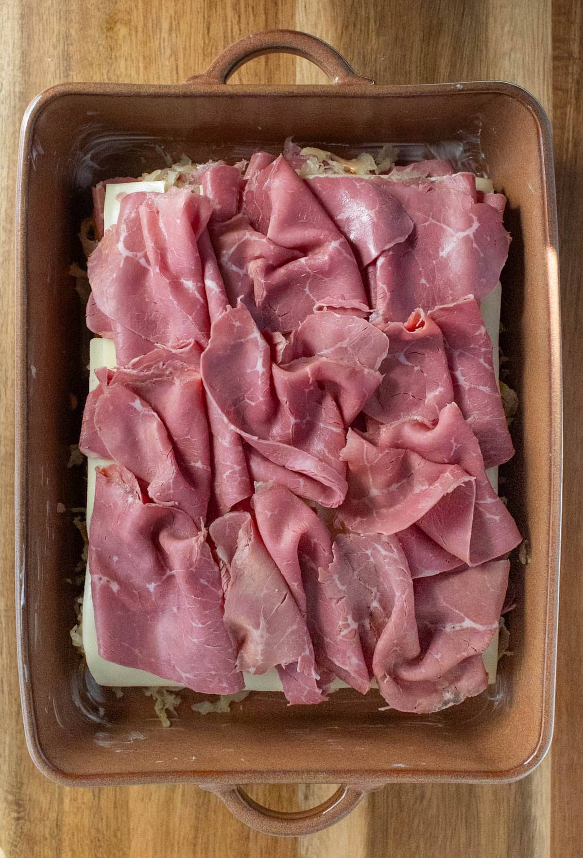 Sliced corned beef on top of sauerkraut and Swiss cheese on bottoms of rolls in a baking dish.