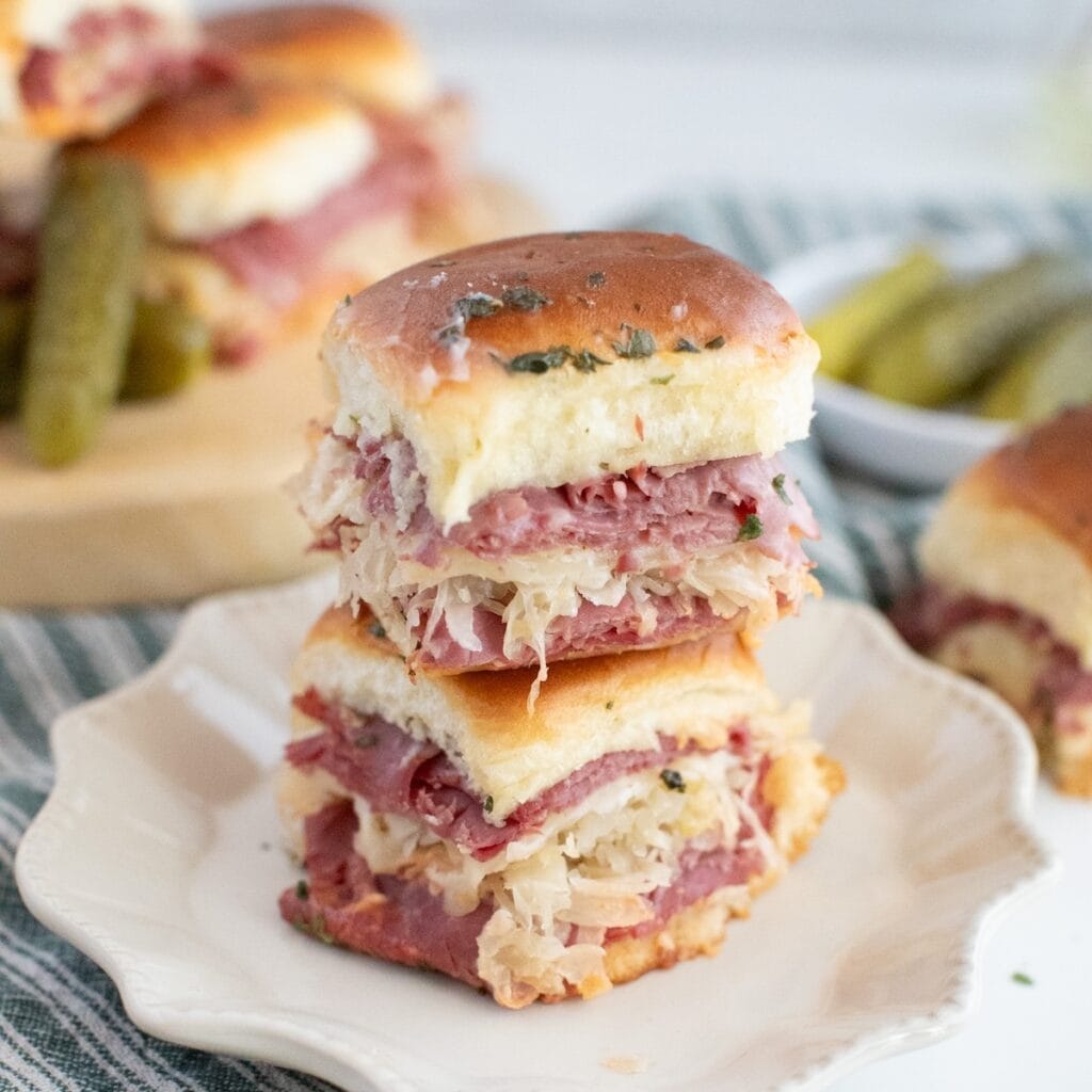 Small Reuben sandwiches on white plate with striped dish towel and more sandwiches in background.