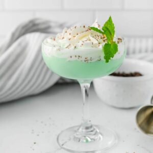 Green cocktail with whipped cream, chocolate shavings, and a sprig of mint on marble counter with striped dish towel.