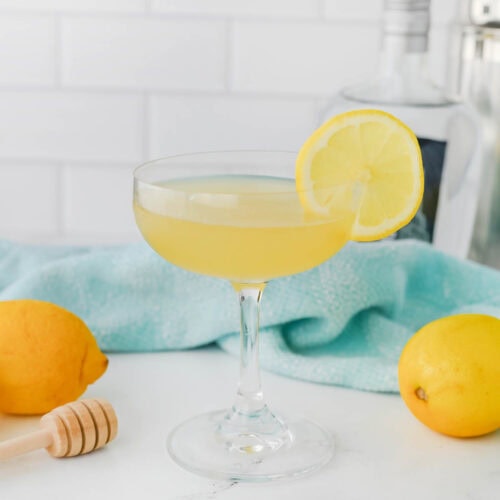 Yellow liquid in a coupe glass with a lemon slice on the rim, a honey dripper, a lemon, and a light blue dishtowel in the background on a white counter.
