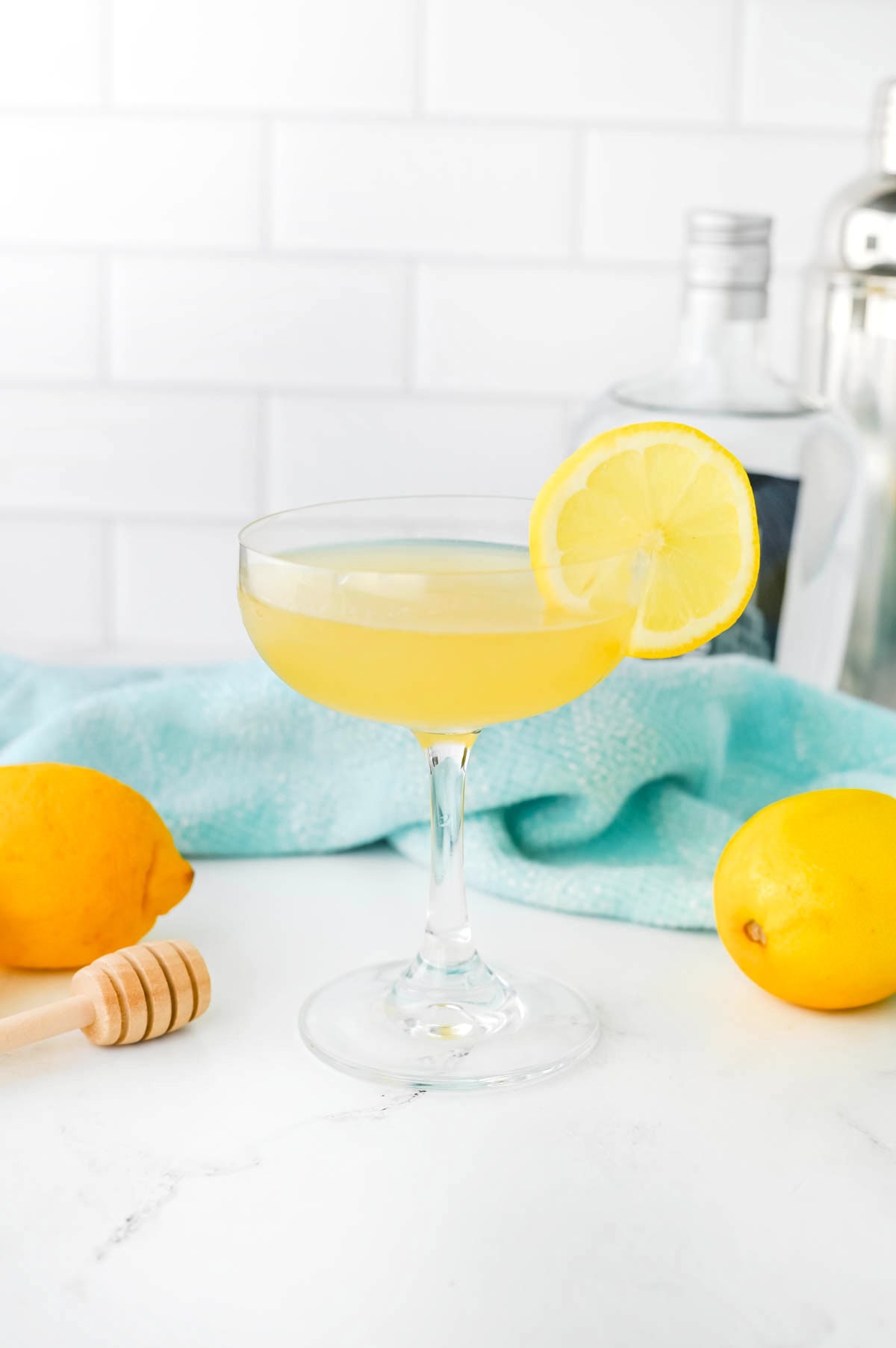 Yellow liquid in a coupe glass with a lemon slice on the rim, a honey dripper, a lemon, and a light blue dishtowel in the background on a white counter.