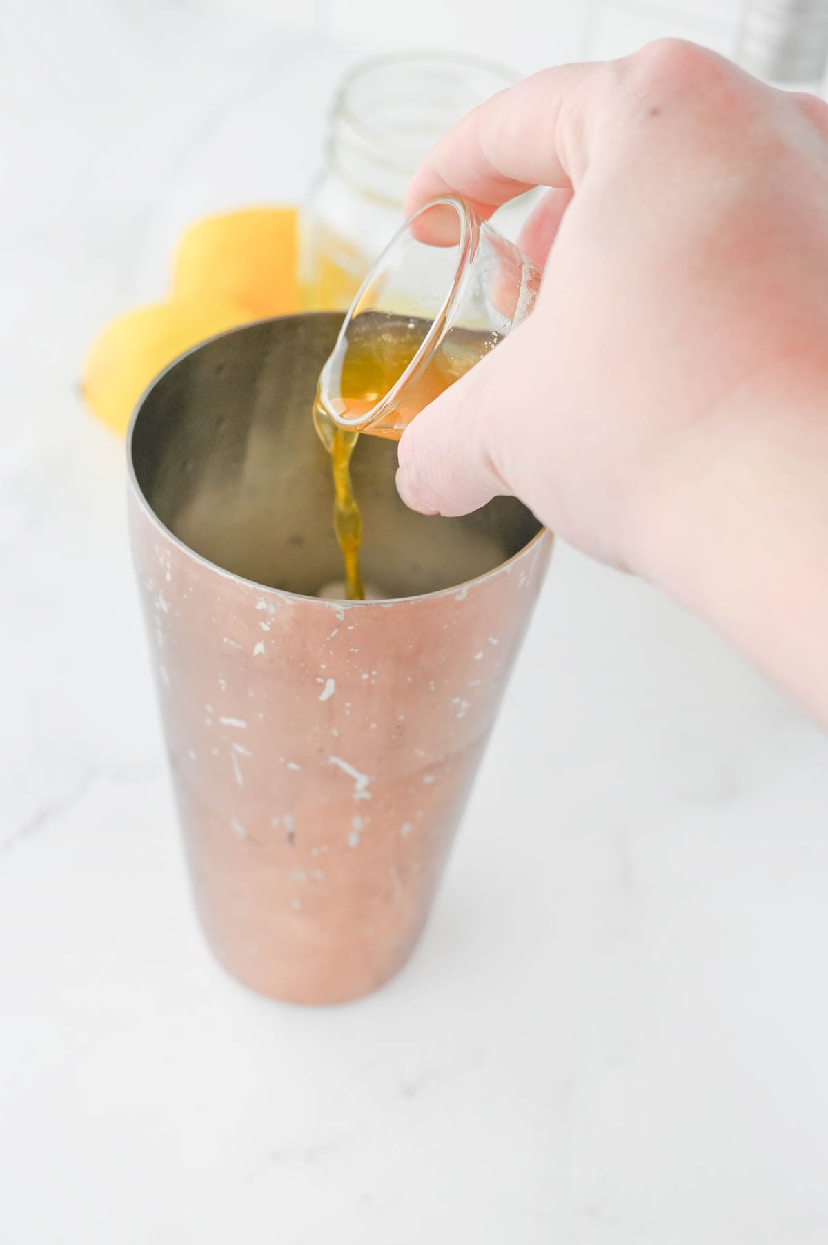 Pouring honey syrup into a cocktail shaker with two lemons in background.