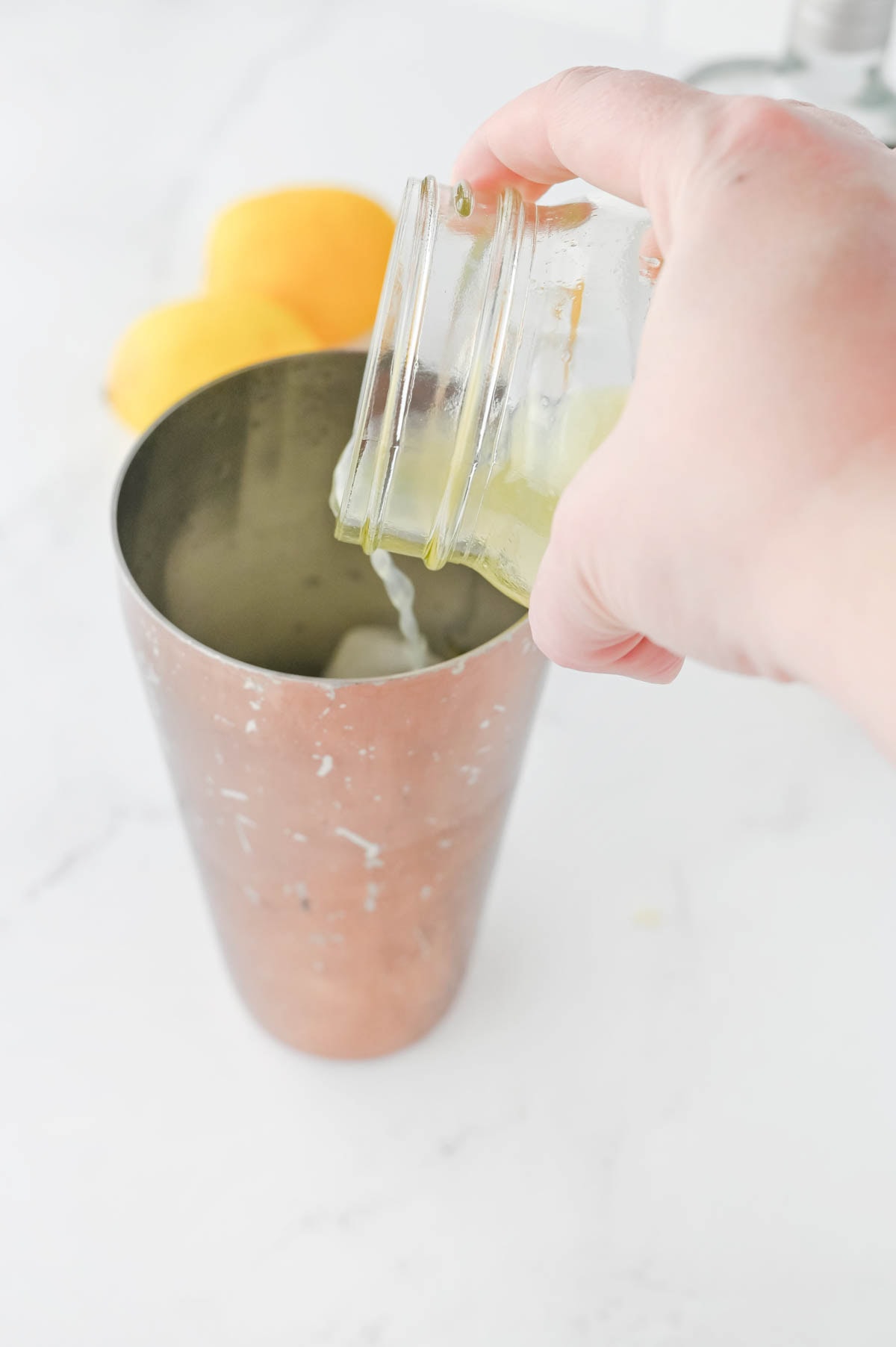 Pouring lemon juice into a cocktail shaker with two lemons in background.