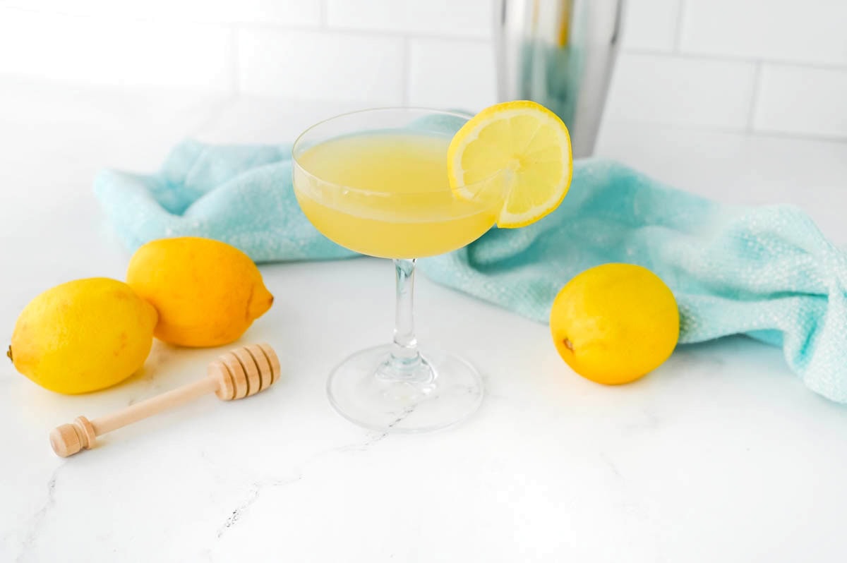 Yellow liquid in a coupe glass with a lemon slice on the rim, a honey dripper, a lemon, and a light blue dishtowel in the background on a white counter.
